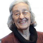 An enhanced color photographic headshot and shoulders cutout of a smiling glasses adorned Amelie O. Rorty with her head tilted to her right facing the camera wearing a black cowl neck under a autumn colored jacket used to visually identify her.