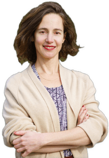 An enhanced color photographic headshot and torso cutout of a mildly smiling standing Anat Schectman facing viewer with arms crossed at waist wearing a purple patterned dress under a beige sweater used to visually identify her.