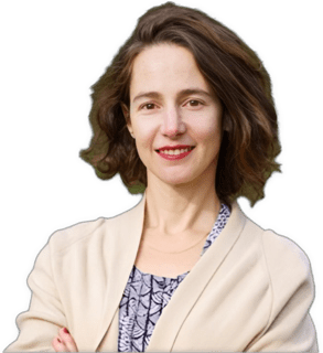An enhanced color photographic headshot and upper torso cutout of a mildly smiling standing Anat Schectman facing viewer with arms crossed at waist (barely visible) wearing a purple patterned dress under a beige sweater used to visually identify her.