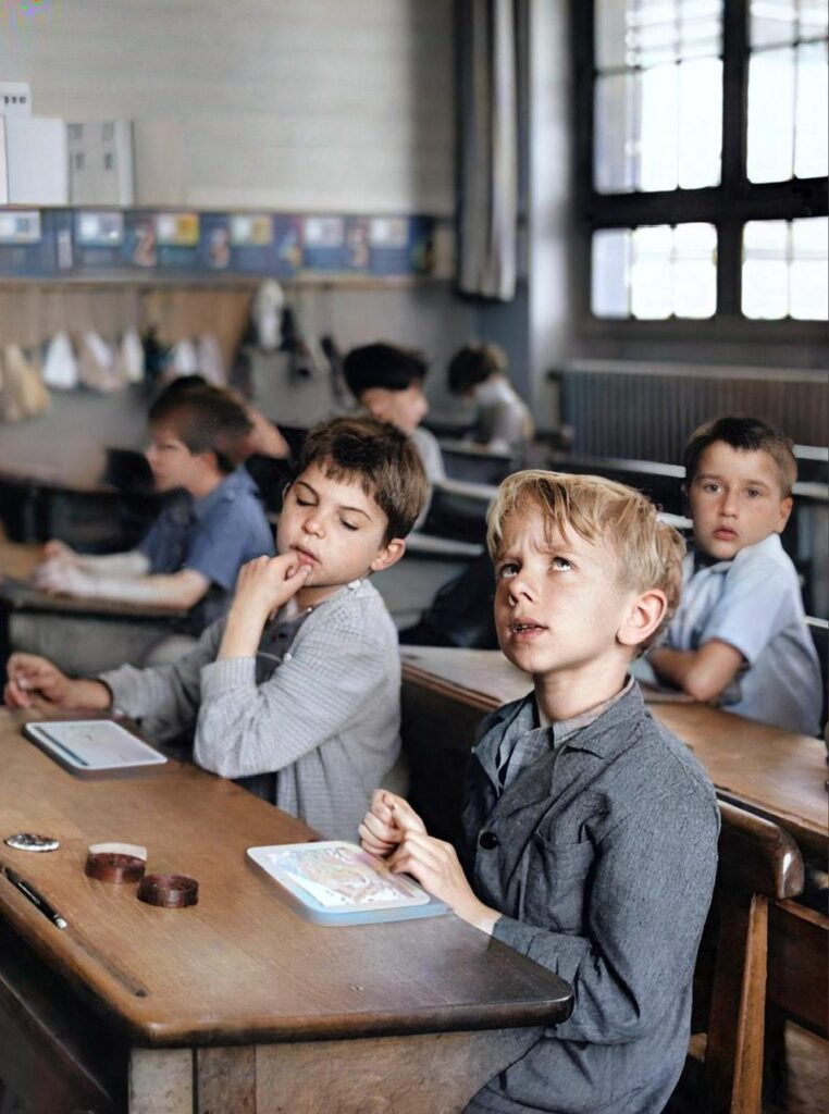 An enhanced colorized photograph of young male schoolchildren in a classroom at their desks with the most prominent boy looking up in thought while the boy to his right is looking at the thinking boy's tablet.