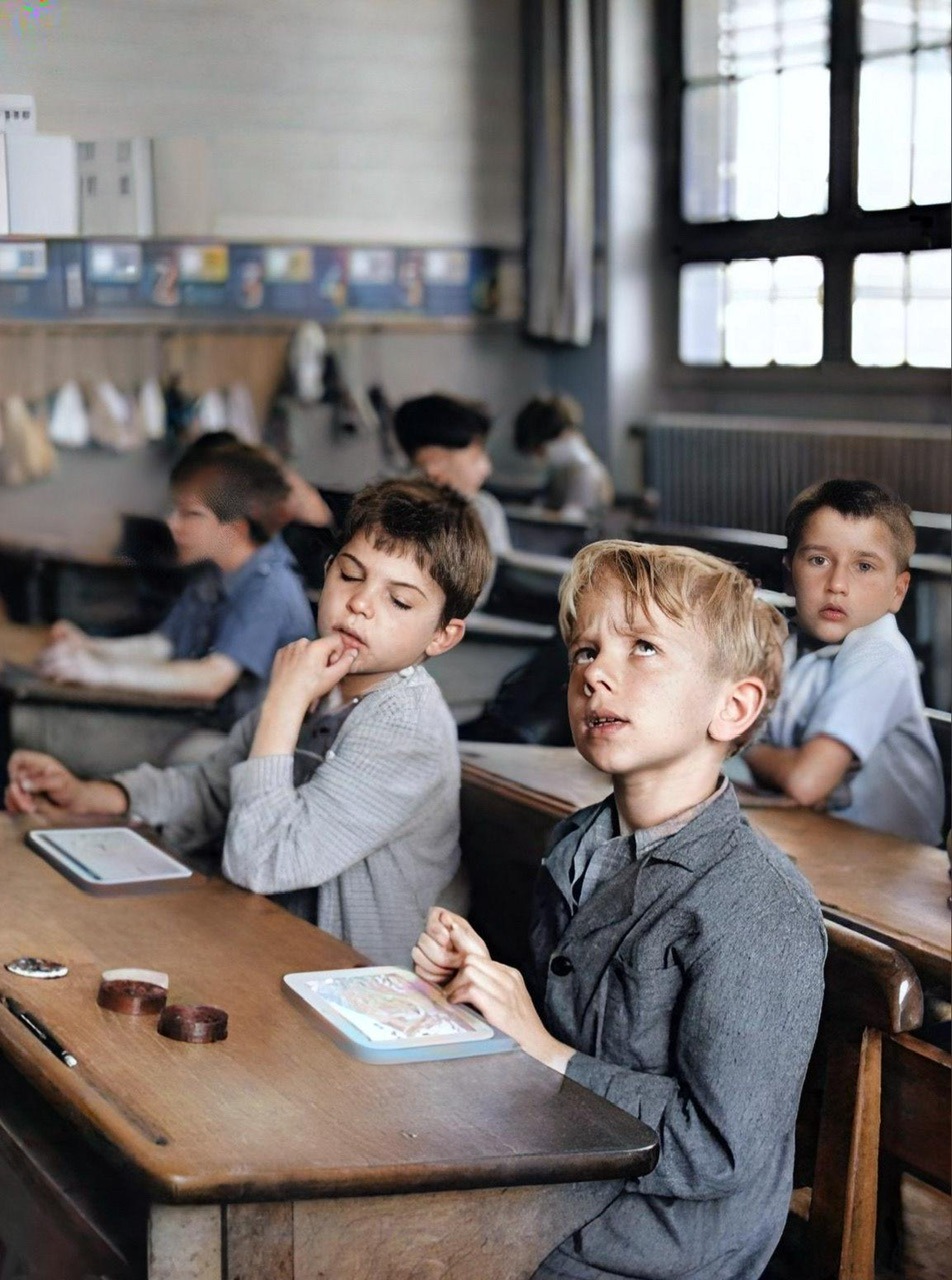 An enhanced colorized photograph of young male schoolchildren in a classroom at their desks with the most prominent boy looking up in thought while the boy to his right is looking at the thinking boy's tablet.