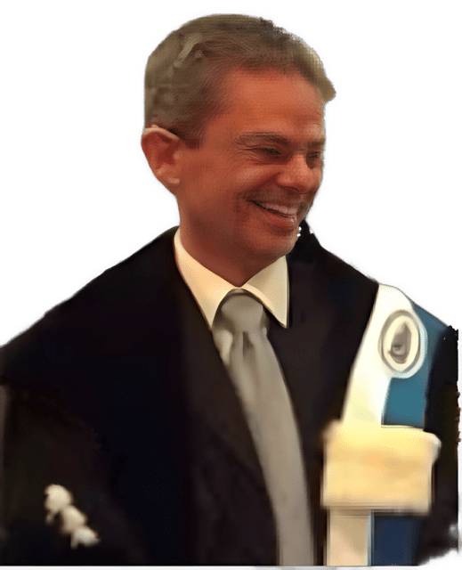 An enhanced color photographic upper torso and headshot cutout of a smiling Dominik Perlerr with rimless glasses with his head bent down looking to his left and wearing a yellow collared shirt with a gray tie under a black suit coat with a blue and white sash over his left shoulder used to visually identify him.