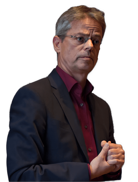 A reversed enhanced color photographic upper torso to headshot cutout of an unsmiling Dominik Perler with rimless glasses with his shoulders turned to his left while grasping his hands together at his solar plexus while wearing a dark maroon open collared shirt with no tie under a black suit coat used to visually identify him.
