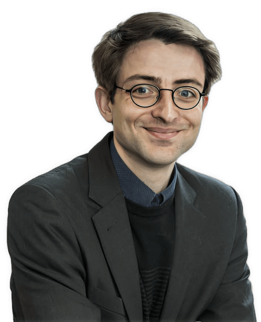 A reversed, enhanced color photographic headshot cutout of Kurt Sylvan wearing glasses, a closed-mouth smirk, and a dark blue shirt under a black sweater under a dark suit coat is used for visual identification.