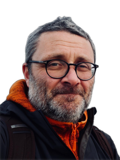 A color photographic cutout of glasses-wearing, salt-and-pepper-bearded Markus Wild wearing an outerwear double-layered jacket with an orange inner layer is used for visual identification. 